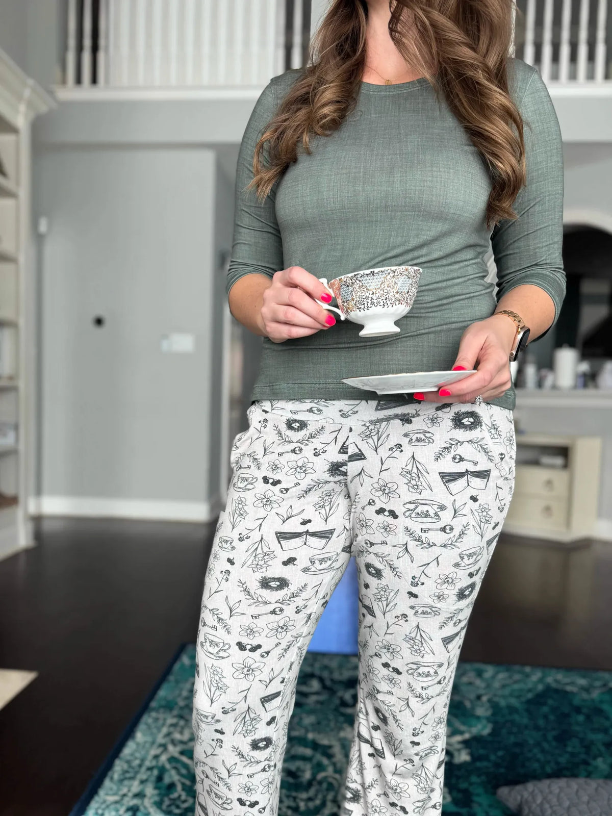 A woman in a green shirt holds a teacup, wearing pants with a tea and toile pattern.