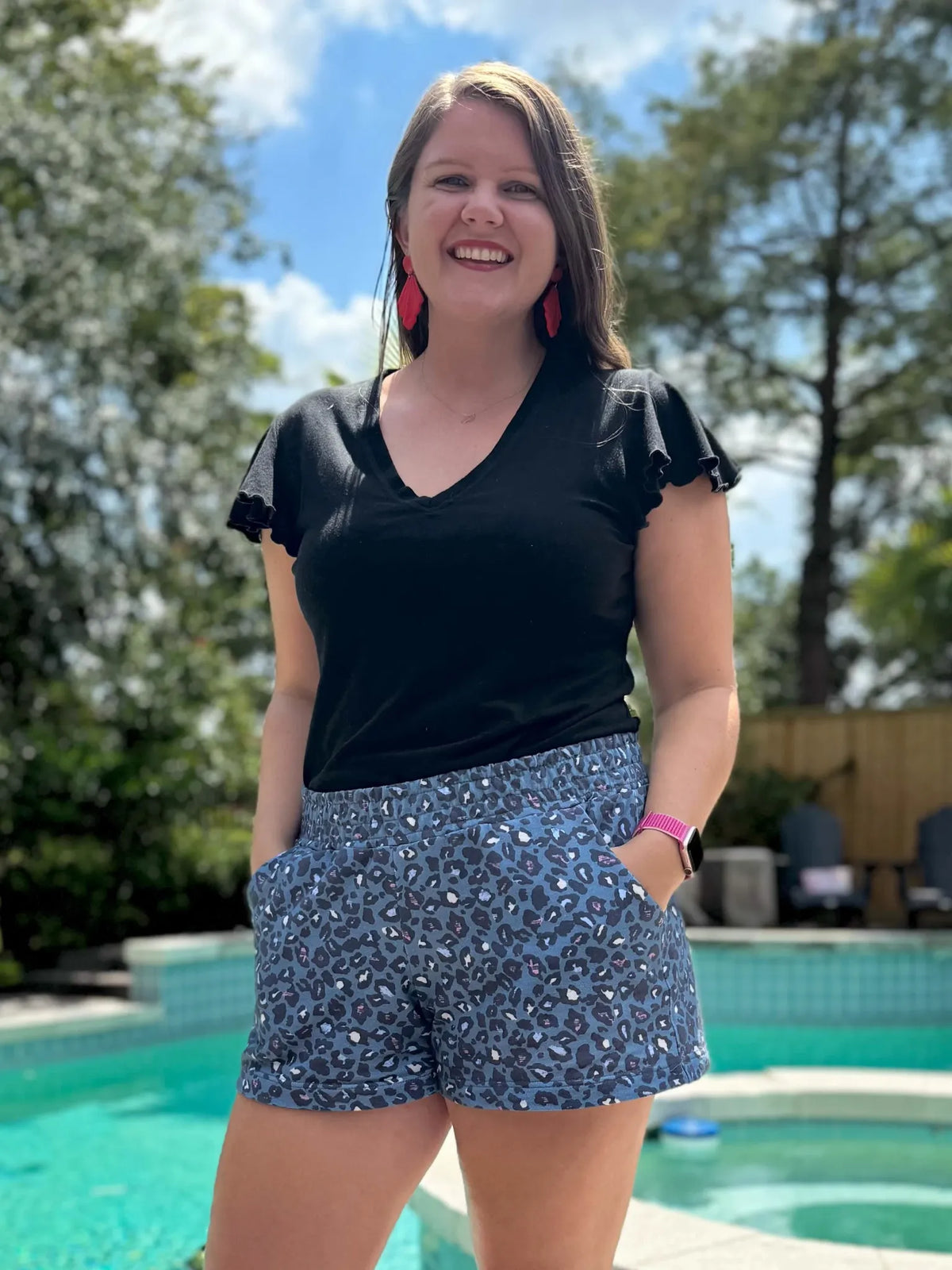 A woman smiles, wearing a black shirt and blue leopard print shorts by a pool.