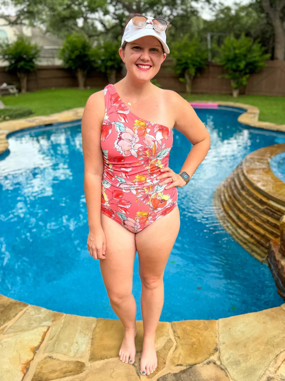 A woman in a coral floral one-shoulder swimsuit stands by a pool.
