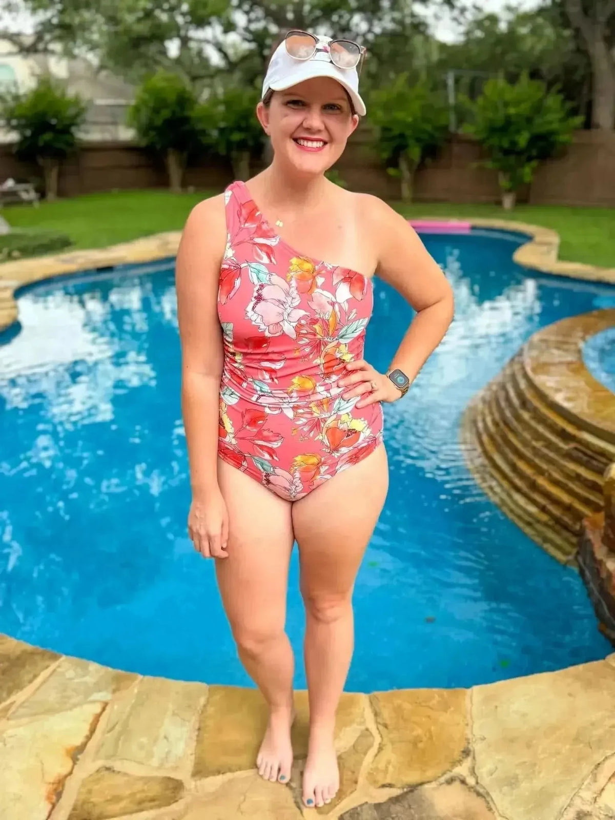 Woman modeling a one-shoulder floral swimsuit in coral pink by the pool.
