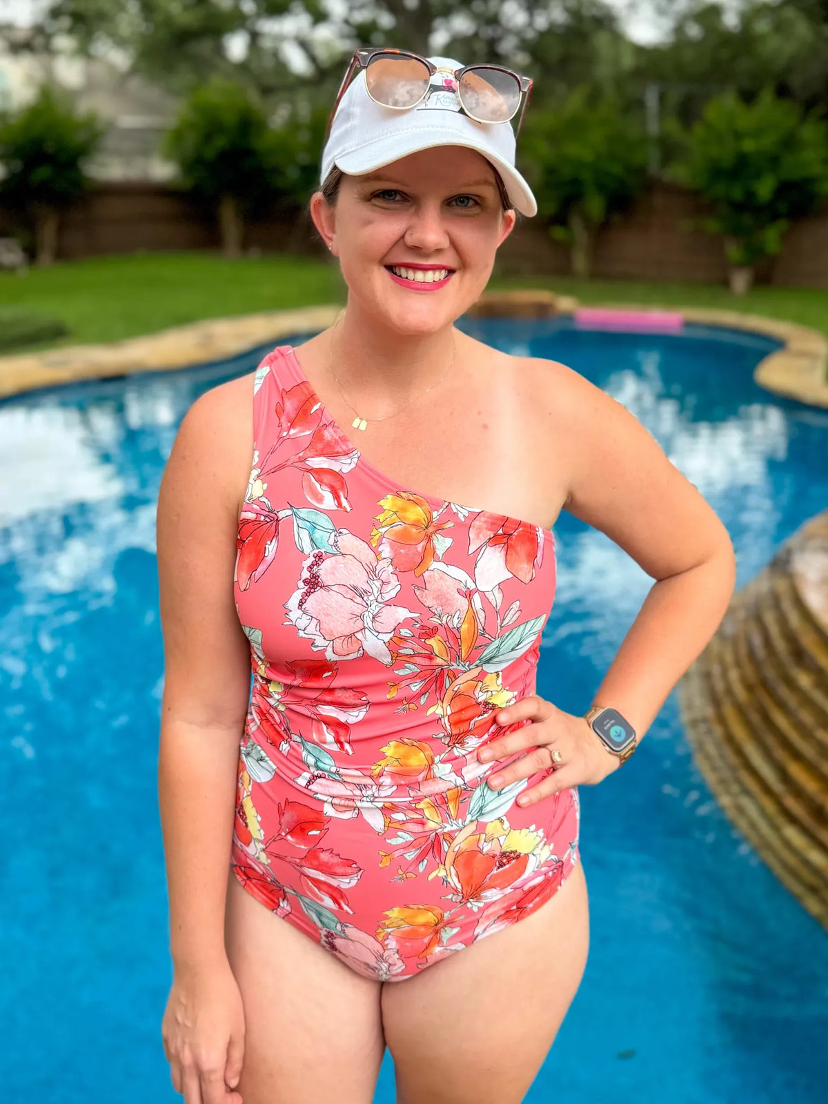 A woman in a coral floral one-shoulder swimsuit stands by a pool.