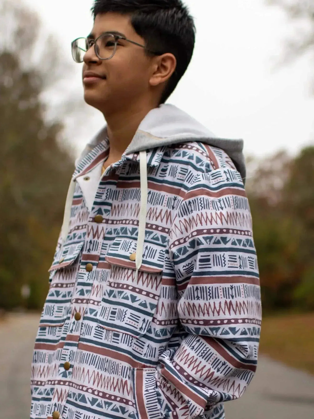Young man wearing a gray hoodie over a patterned navy and rust jacket outdoors.
