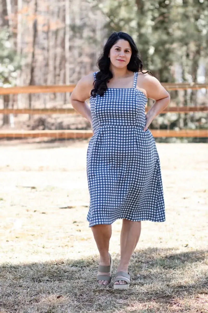 A woman in a navy gingham dress stands outdoors with hands on her hips.