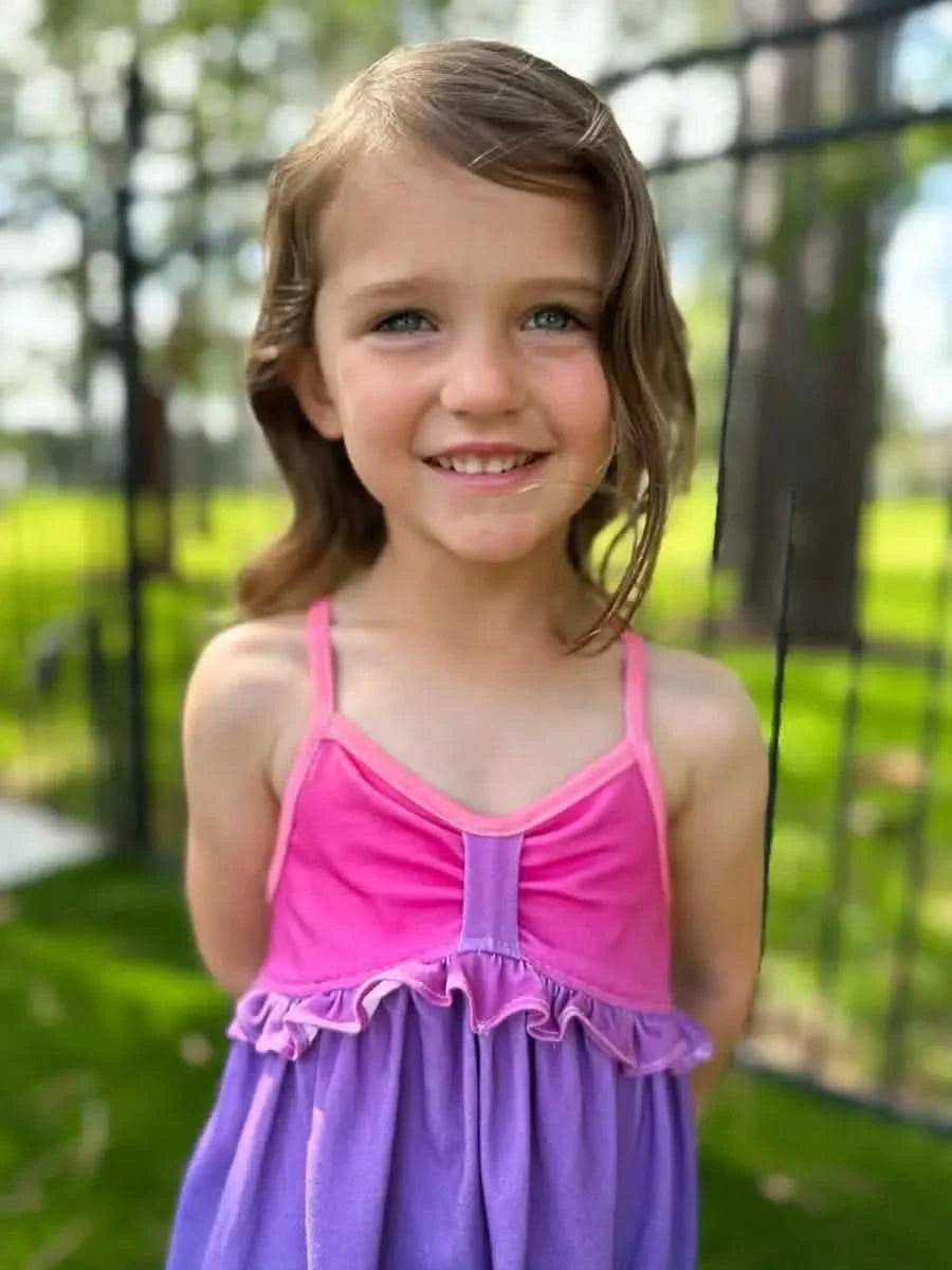 A young girl smiles, wearing a pink and purple ombre dress with ruffles.