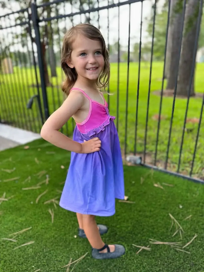 A young girl in a pink and purple ombre dress stands on green artificial grass.