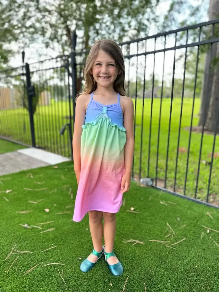 A young girl smiles, wearing a blue, green, and pink ombre dress with teal shoes.