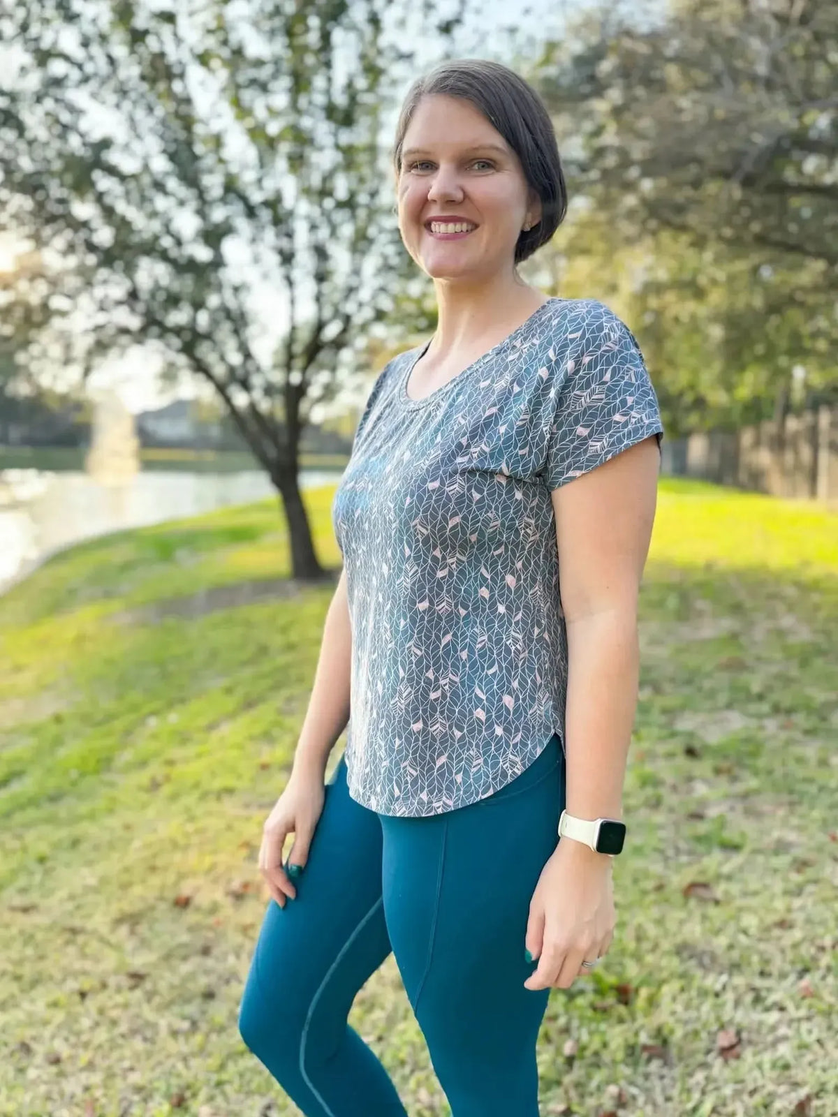 A woman smiles, wearing a blue top with a gold leaf pattern and blue leggings.