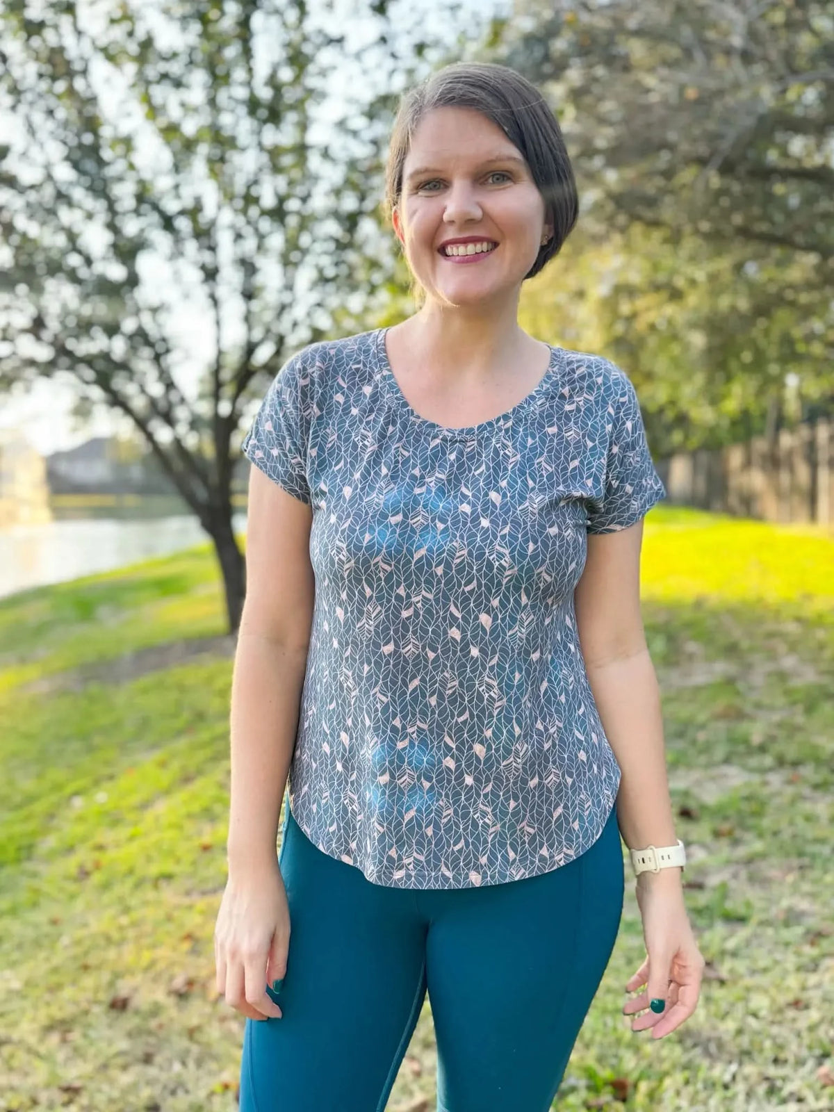 A woman smiles wearing a blue shirt with a gold leaf pattern and teal leggings.