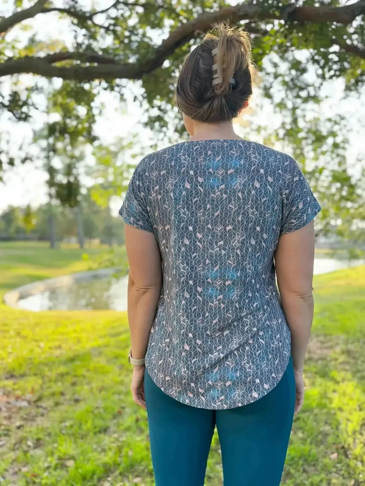 Rear view of a woman wearing a gray short-sleeve top with gold leaf print, outdoors by a pond.