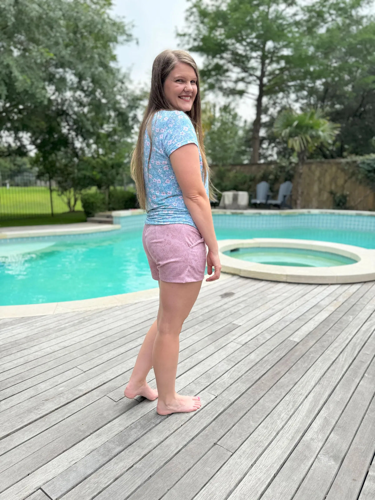 A woman in a mauve stenciled floral top and shorts stands by a pool.
