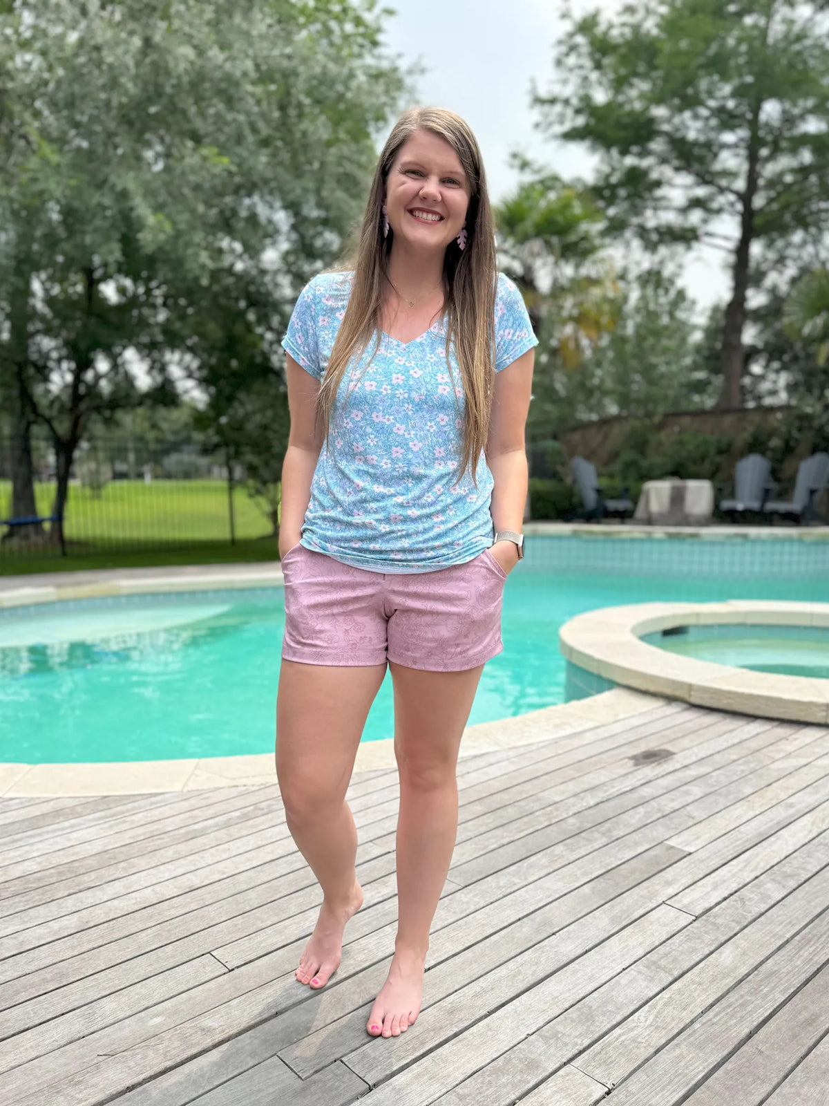 A woman wears a light blue floral top and mauve floral shorts by a pool.