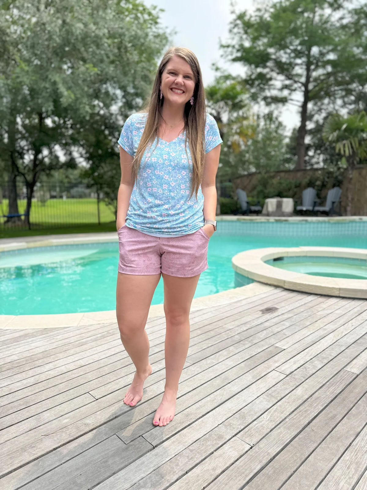 A woman wears a light blue floral top and mauve floral shorts by a pool.
