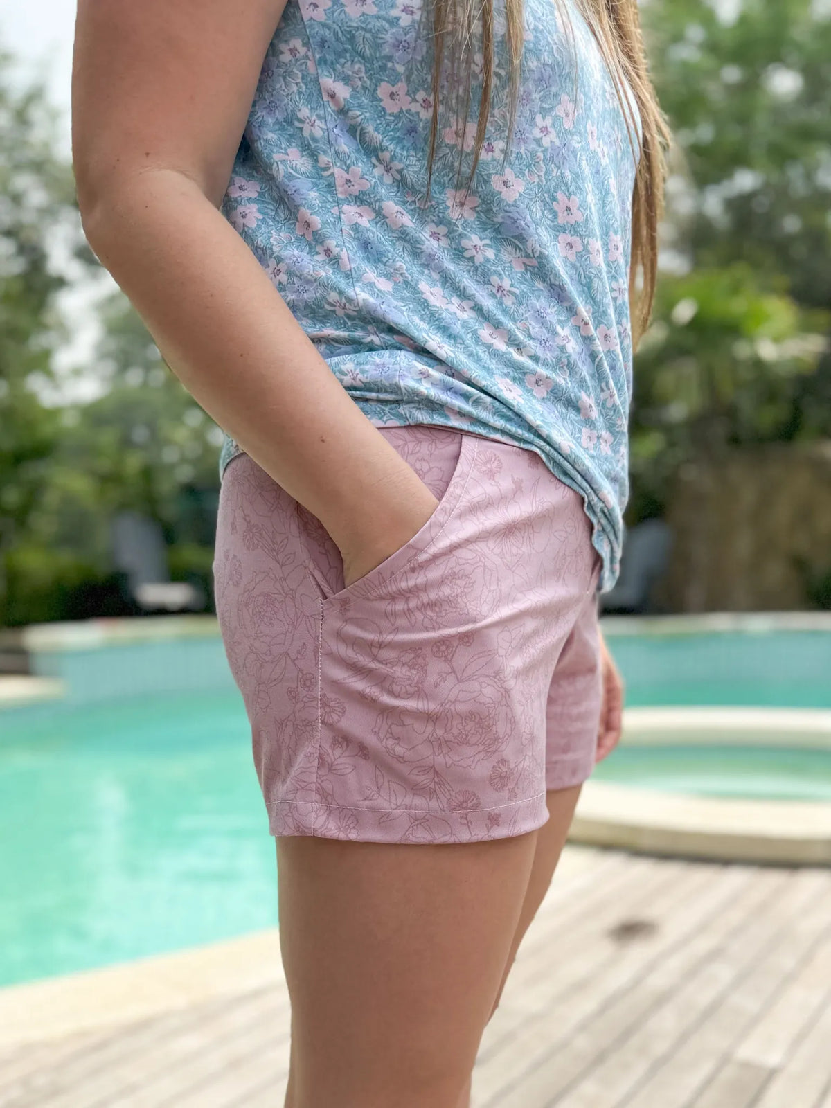 A person wearing a blue floral top and mauve floral shorts with a hand in the pocket.