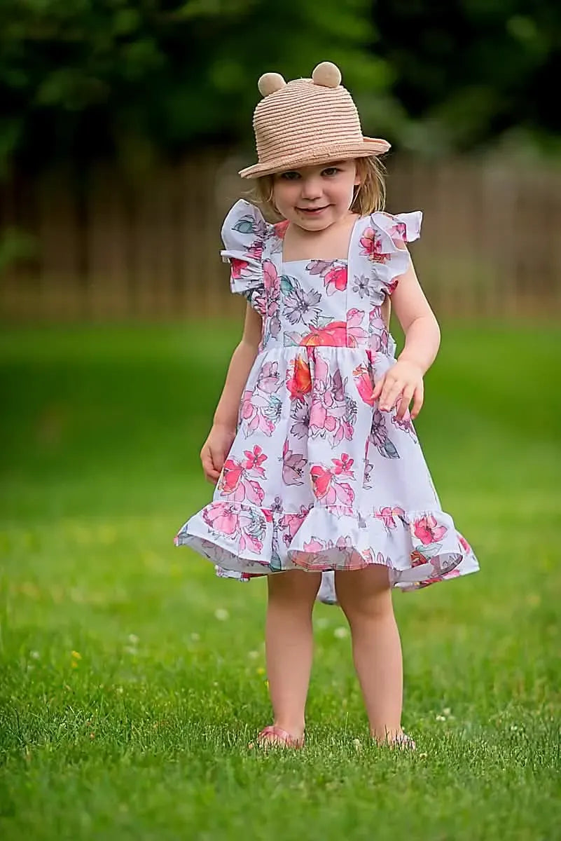 A young girl in a floral dress and straw hat stands in a grassy yard.