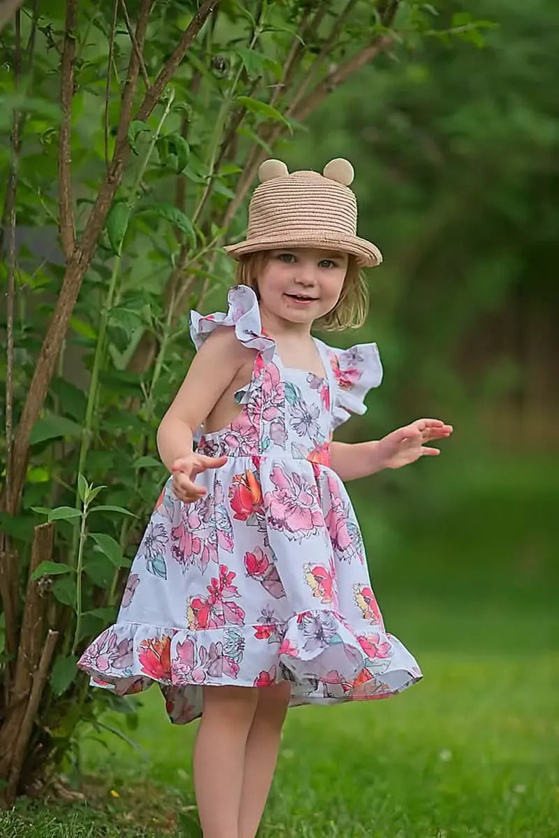 A young girl in a floral dress and straw hat stands in front of green foliage.