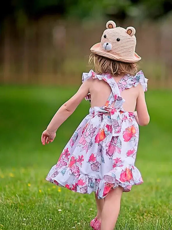A young child in a floral dress and bear hat walks away across a grassy field.