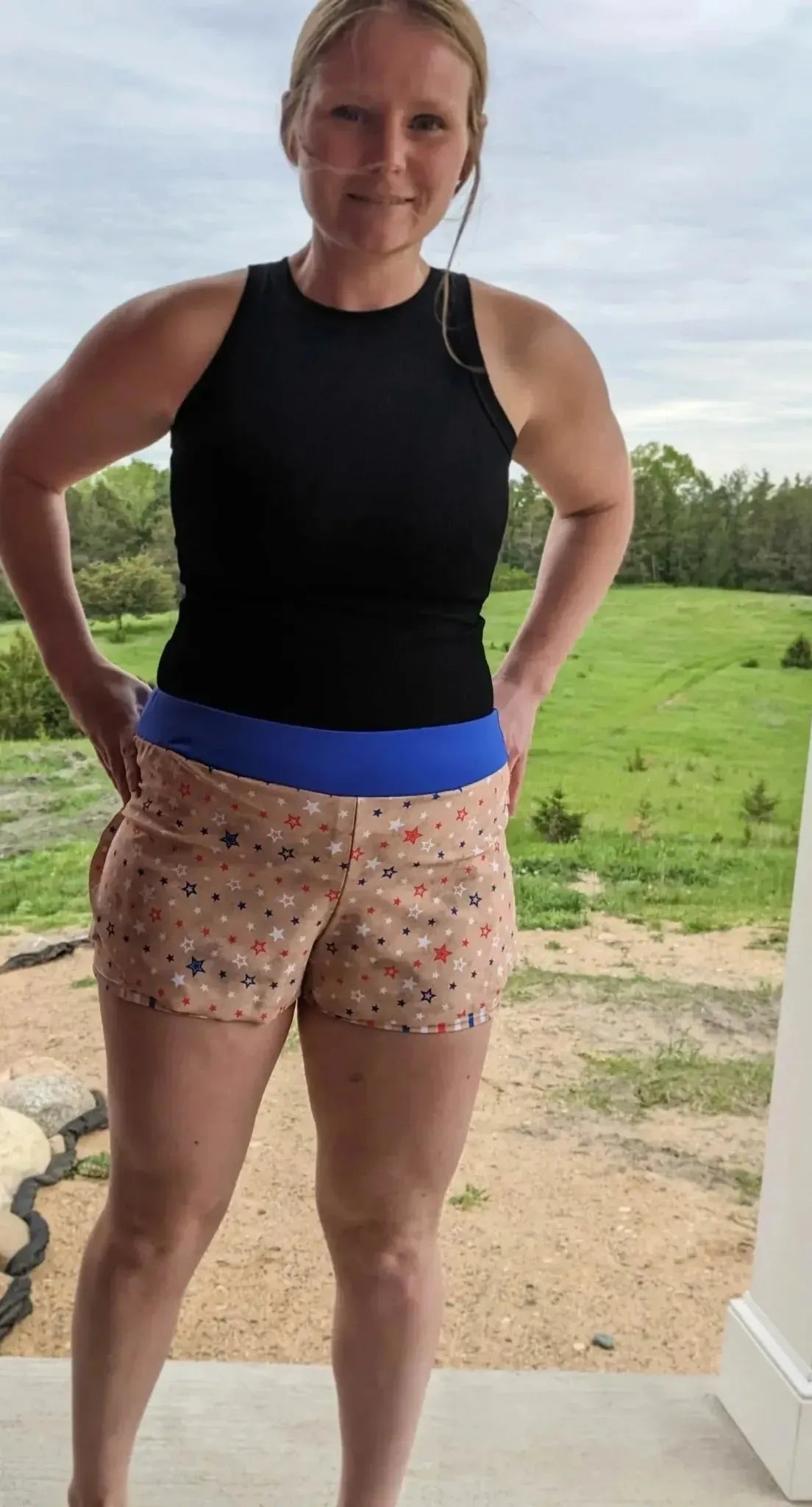 Woman modeling black tank top and orange gingham shorts with blue waistband outdoors.