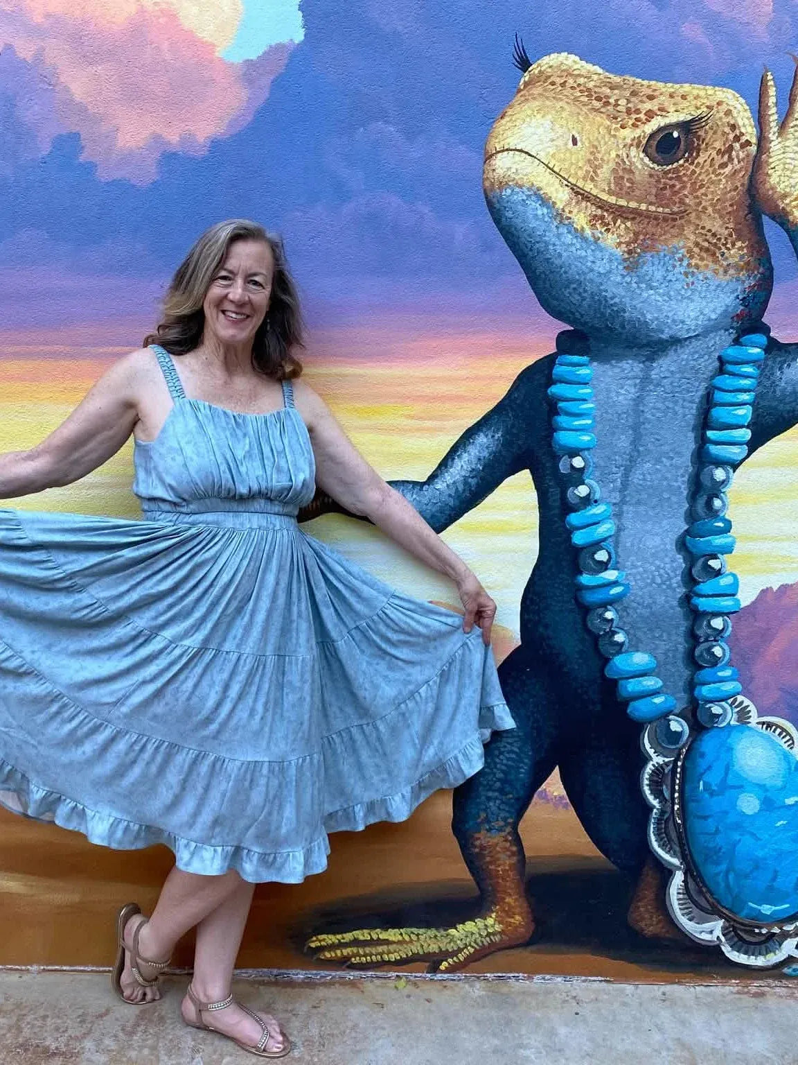 A woman in a dusty blue stenciled floral dress stands next to a lizard mural.