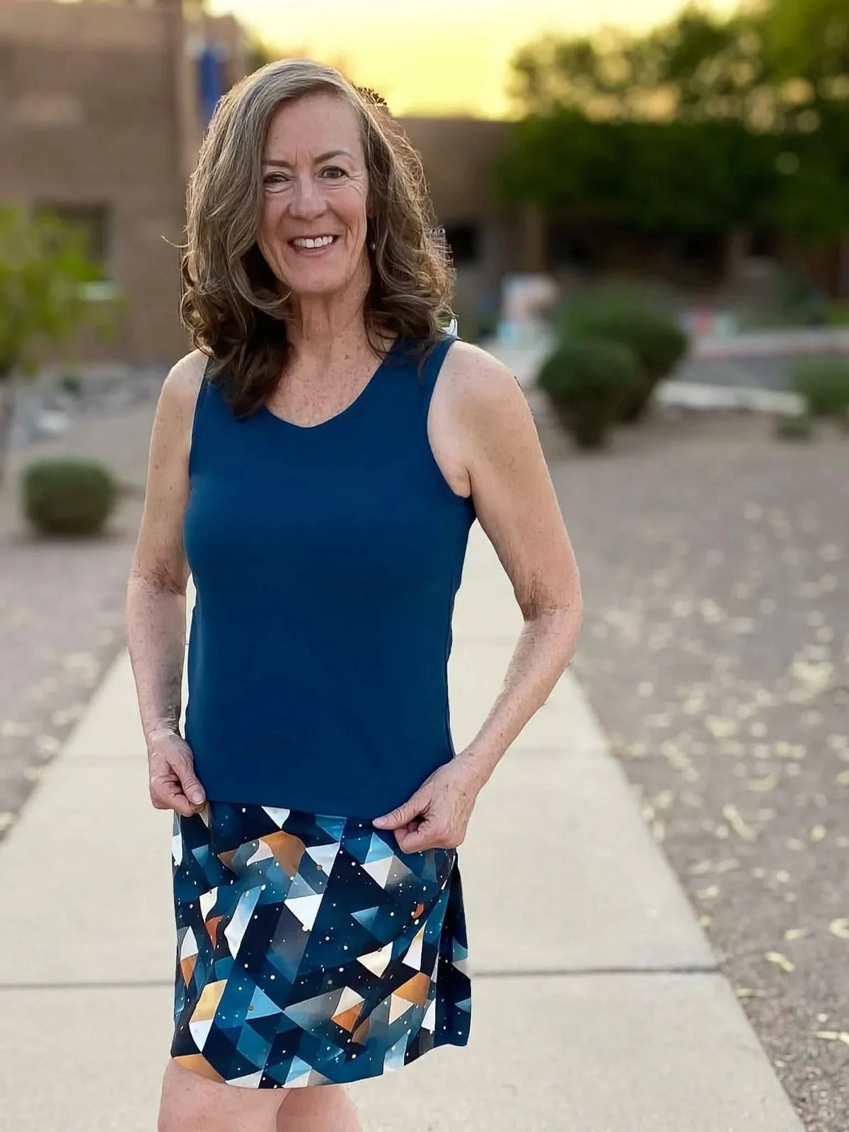 A woman smiles, wearing a teal tank top and a geometric print skirt.