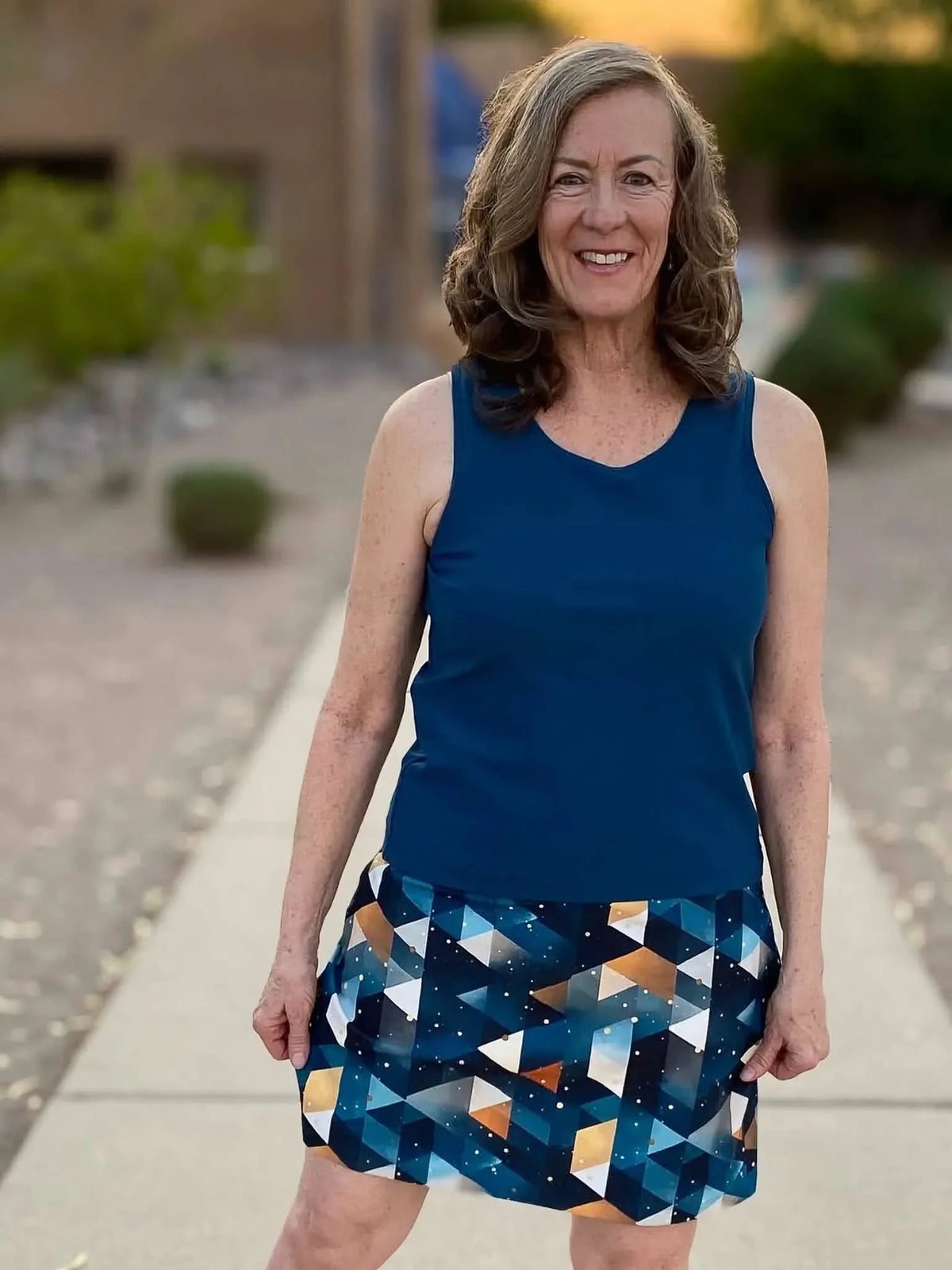 A woman smiles, wearing a teal tank top and a geometric patterned skirt.