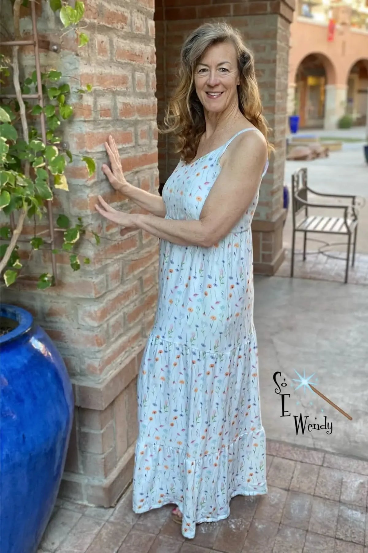 A woman in a white floral dress stands next to a brick pillar.