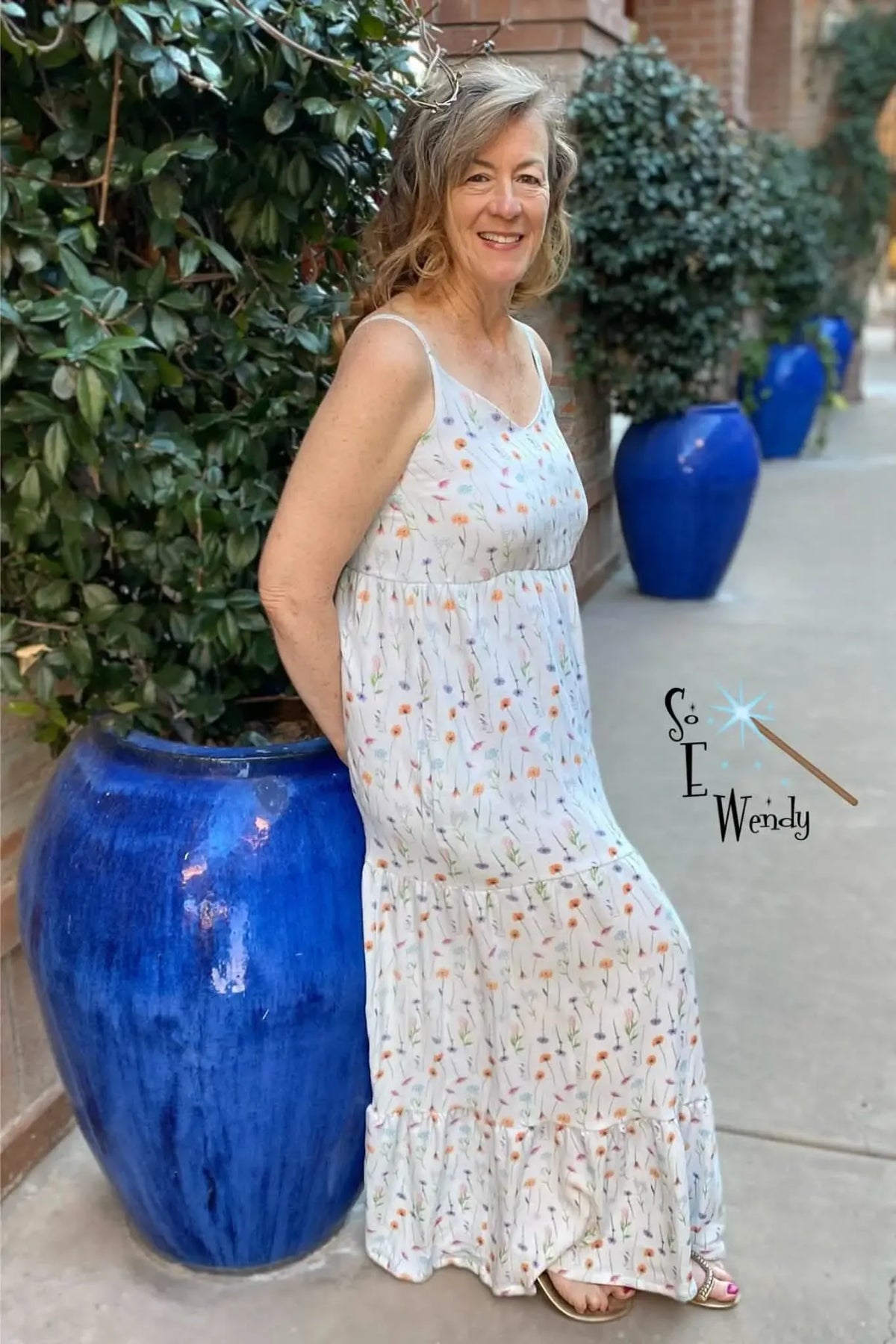 A woman in a white floral dress leans against a blue pot.