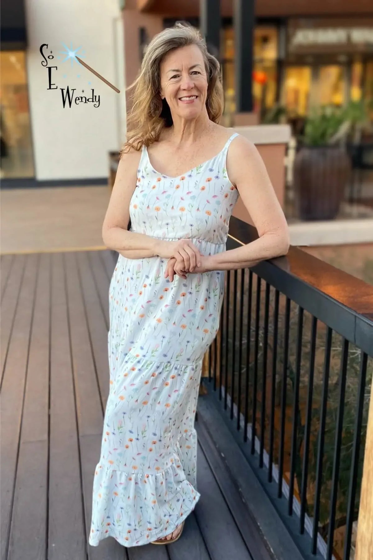 A woman in a floral maxi dress leans on a railing outdoors.