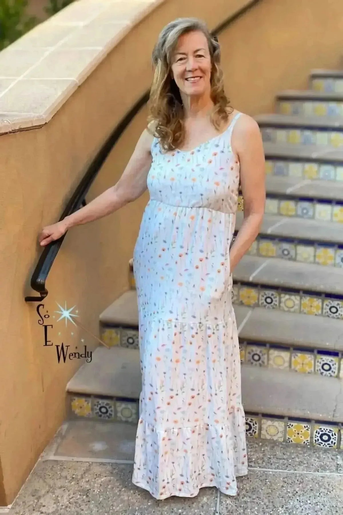 White maxi dress with spaghetti straps, empire waist, tiered skirt, and wildflower print, modeled on outdoor stairs.