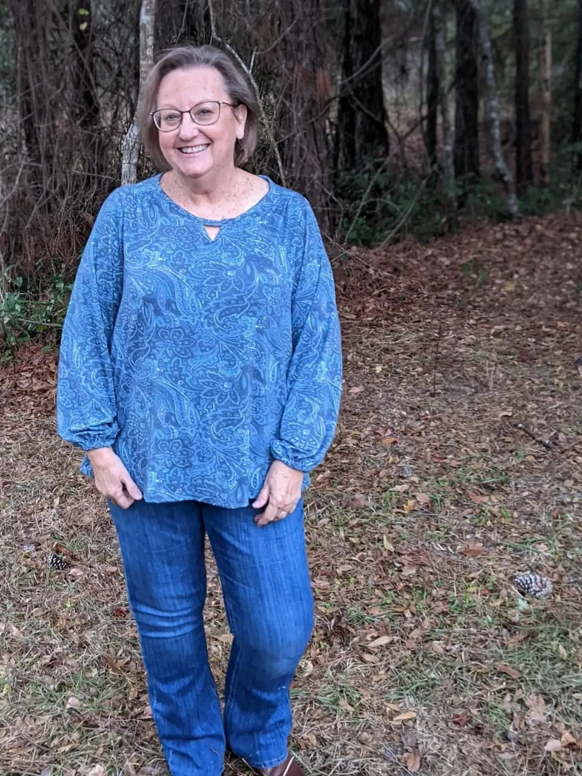 A woman in a blue paisley top and jeans stands in a wooded area.
