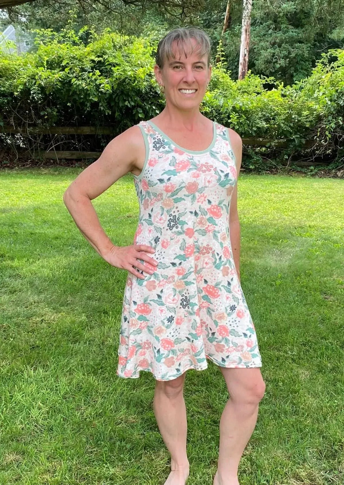 A woman in a floral dress with blush flowers and green trim stands in a grassy yard.