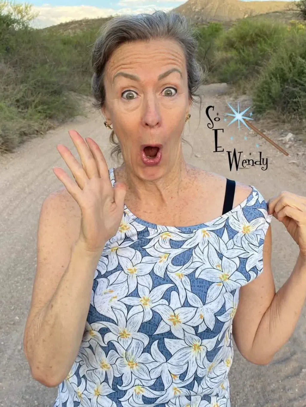 A woman in a blue and white lily print top stands on a dirt road.
