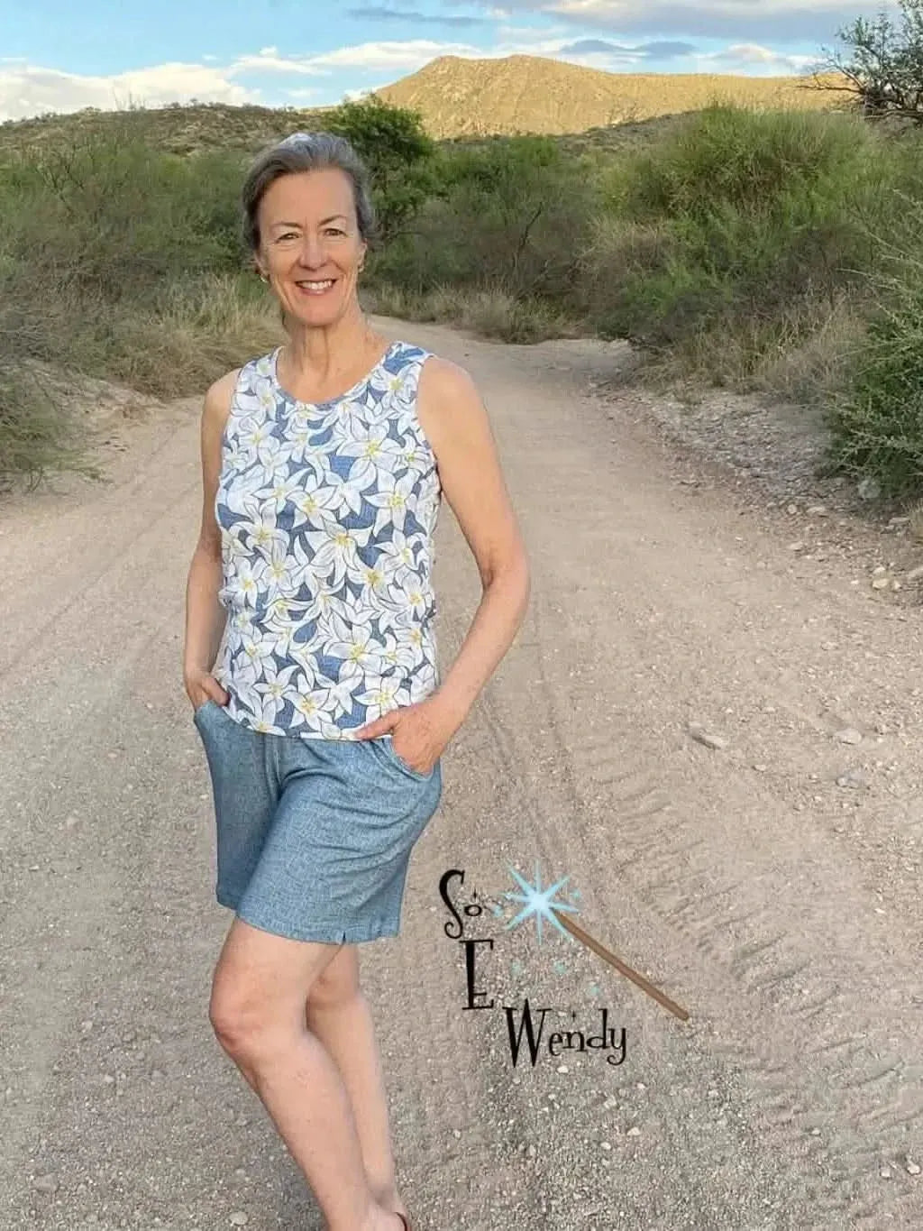 A woman wears a sleeveless top with a blue lilies floral print and light blue shorts.