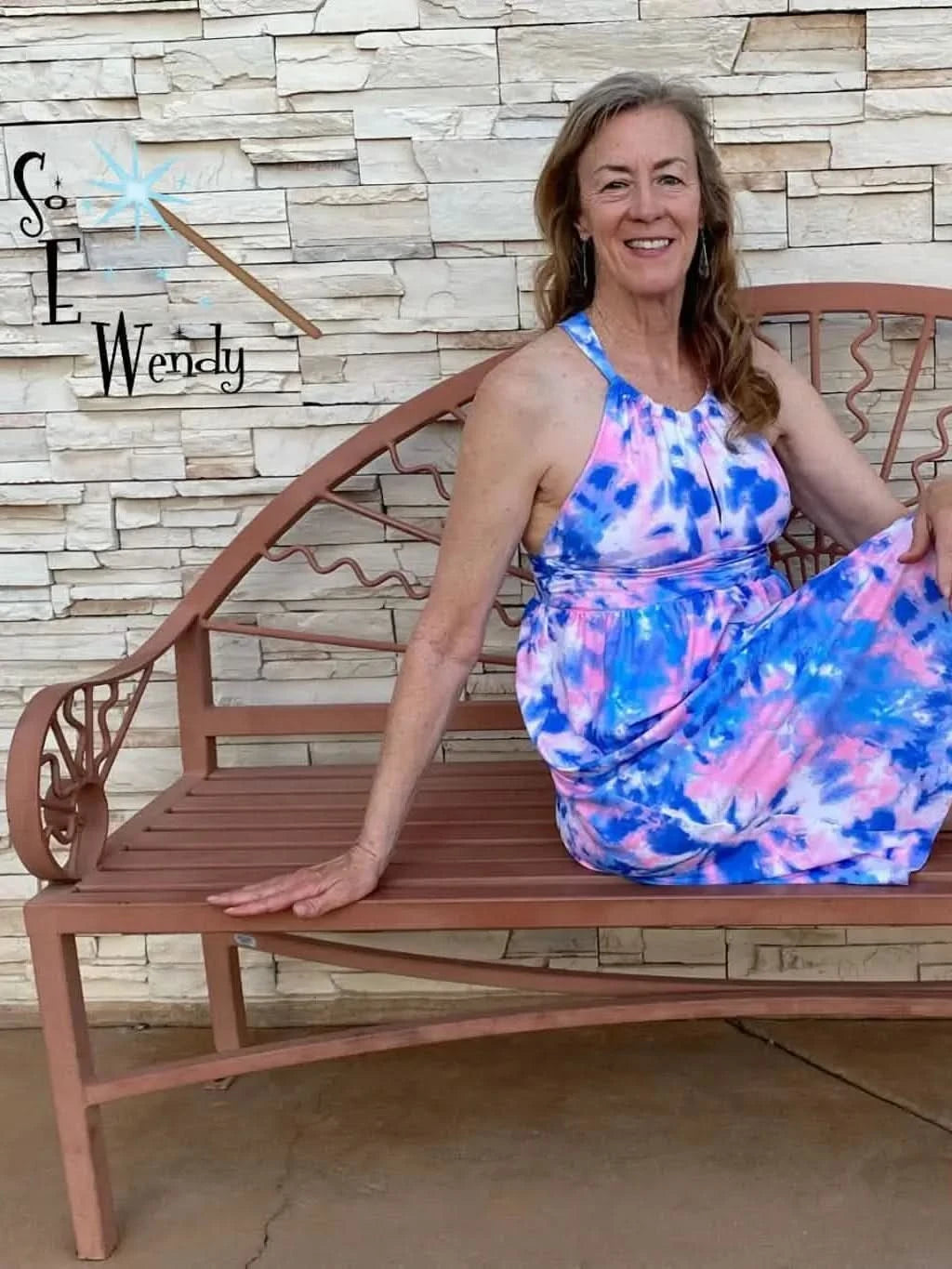 A woman in a pink and blue tie-dye dress sits on a brown metal bench.