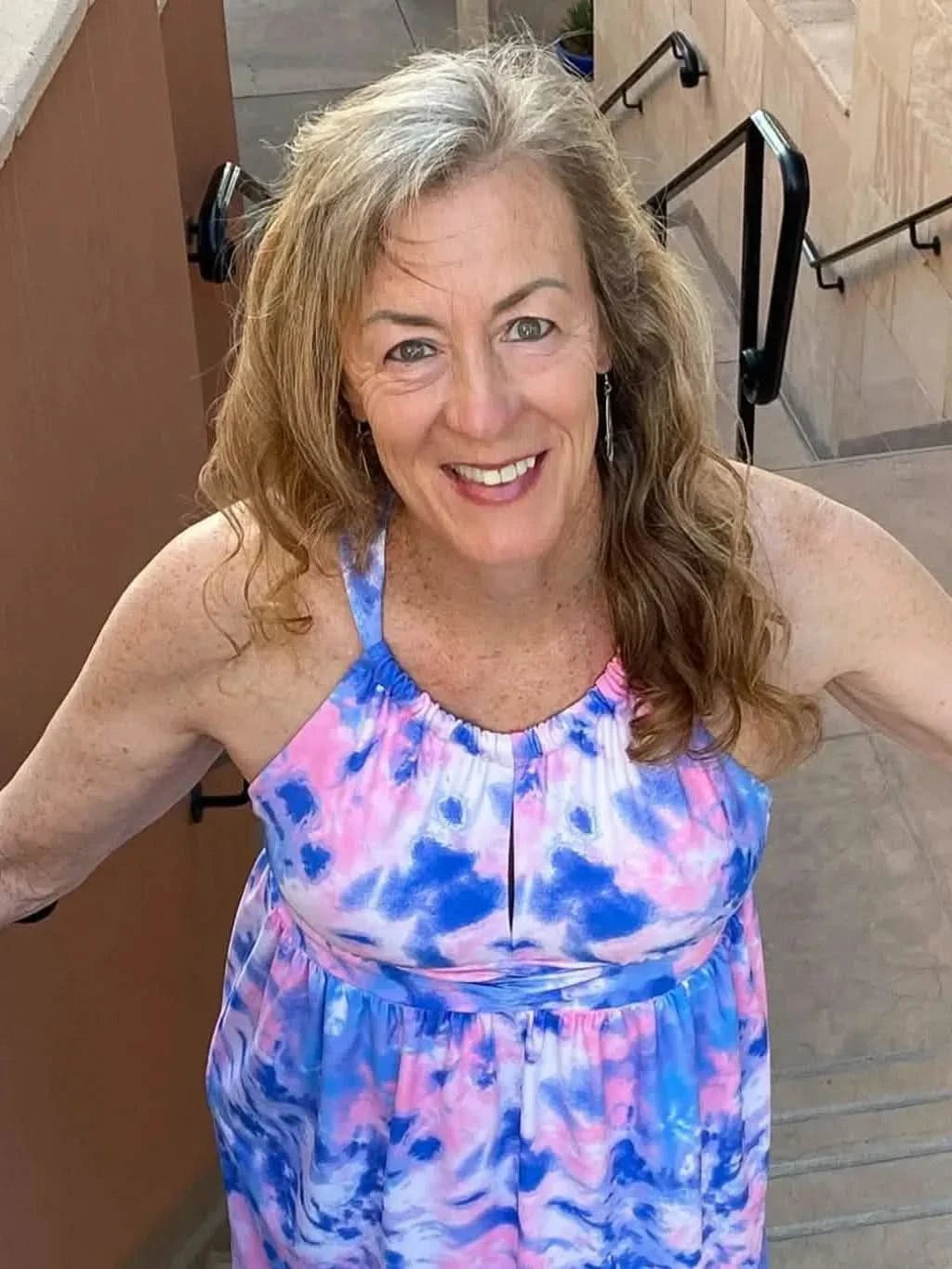 A woman in a pink and blue tie-dye dress smiles while holding onto stair railings.