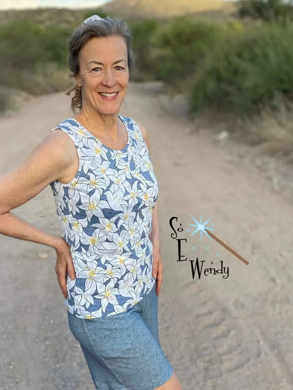A woman smiles, wearing a blue floral tank top and blue shorts on a dirt road.