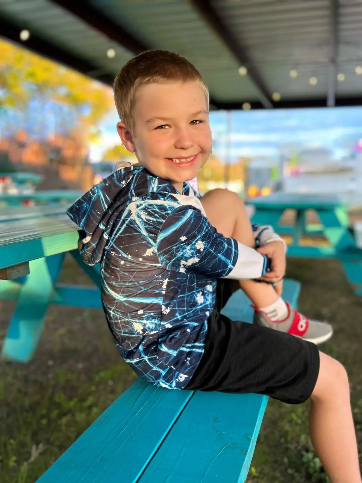 A boy wears a navy cracked ice hoodie while sitting on a teal picnic table.