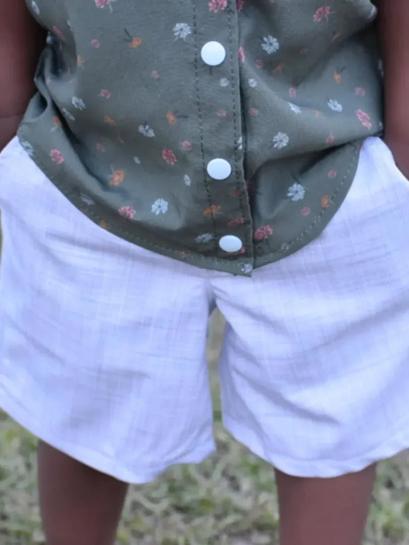 A child wears a green floral top and white plaid shorts with hands in pockets.