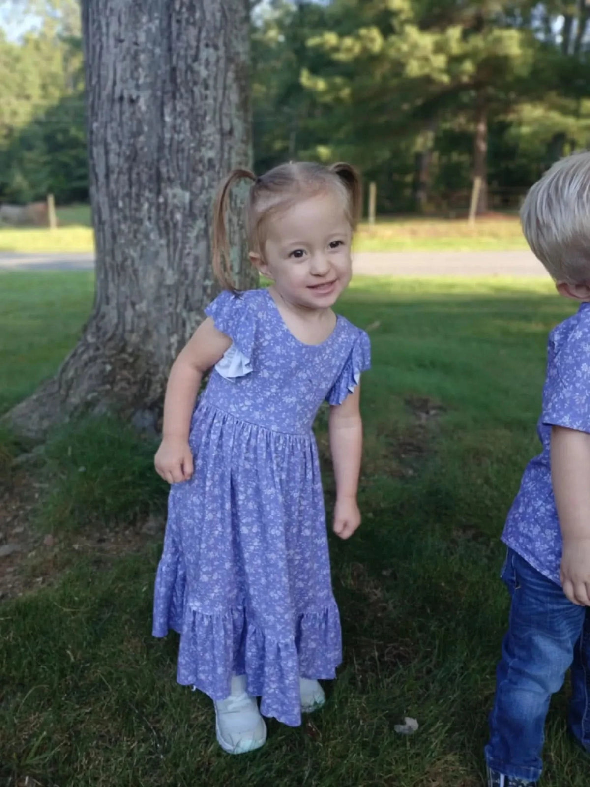 A young girl in a purple floral dress with ruffled sleeves stands next to a tree.