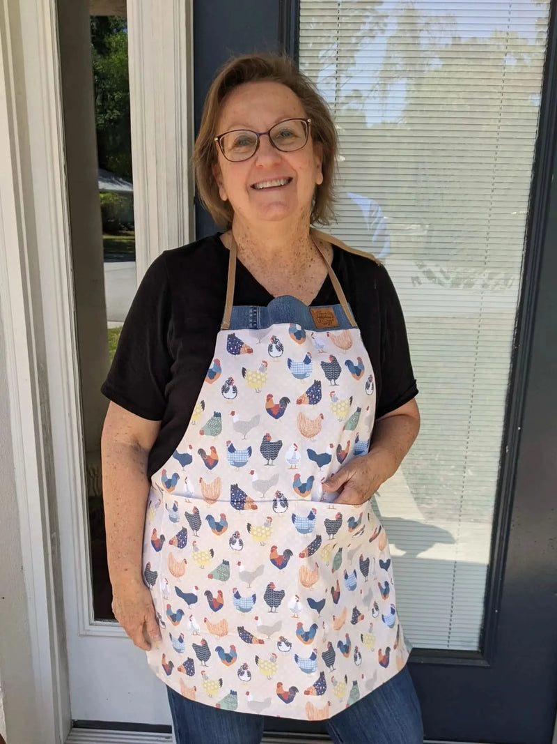 A woman smiles, wearing an apron with a patchwork chicken pattern.