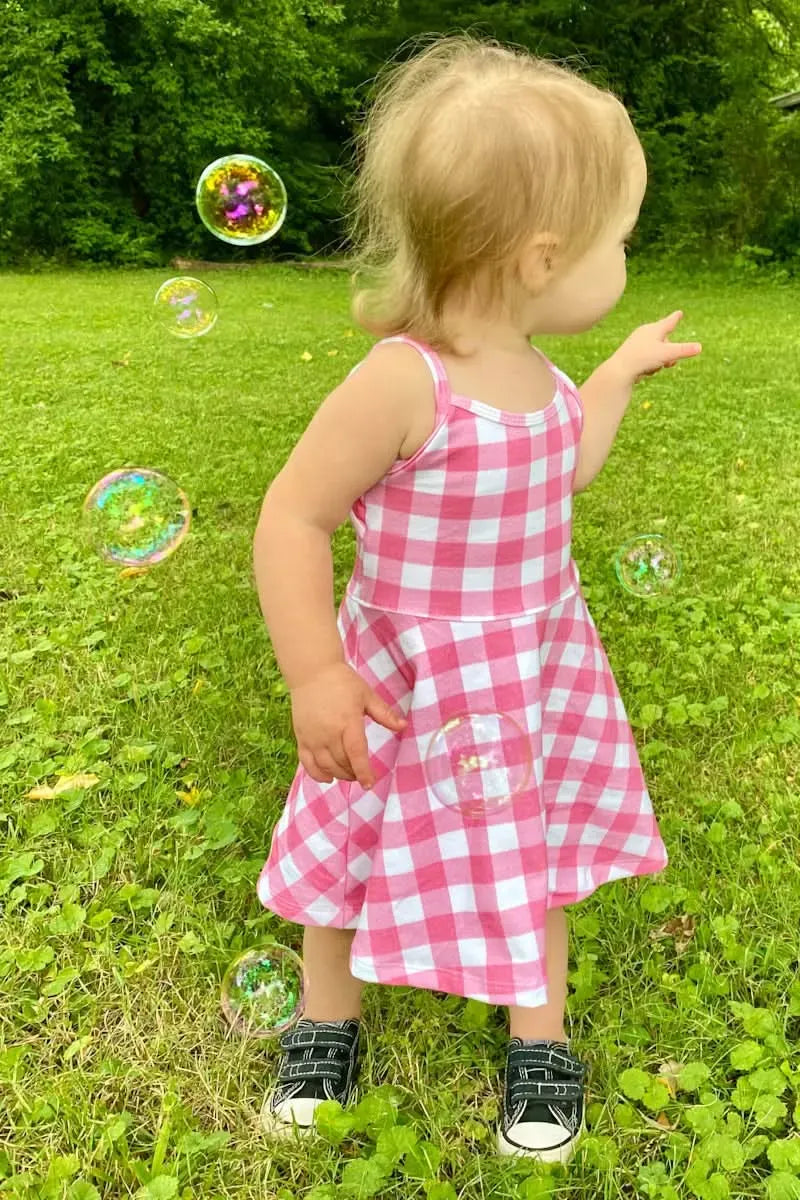 A toddler in a pink and white gingham dress plays with bubbles outdoors.