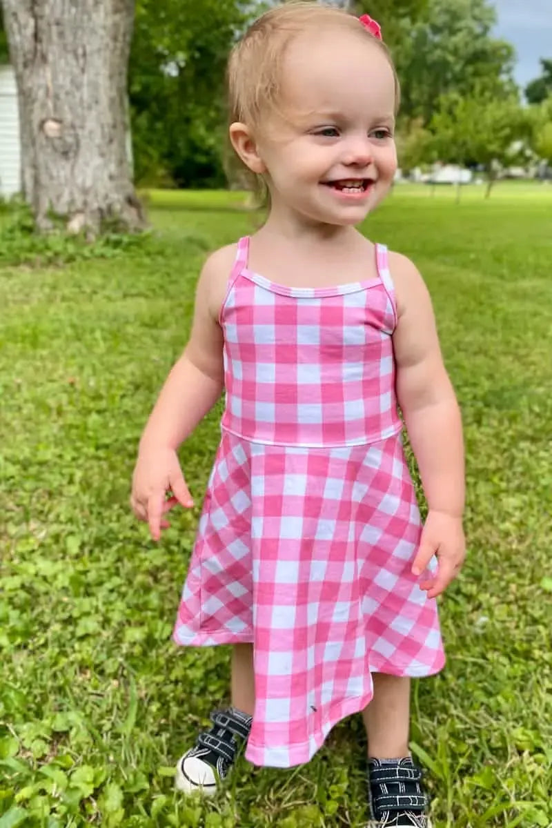 A toddler in a pink and white gingham dress stands on grass.
