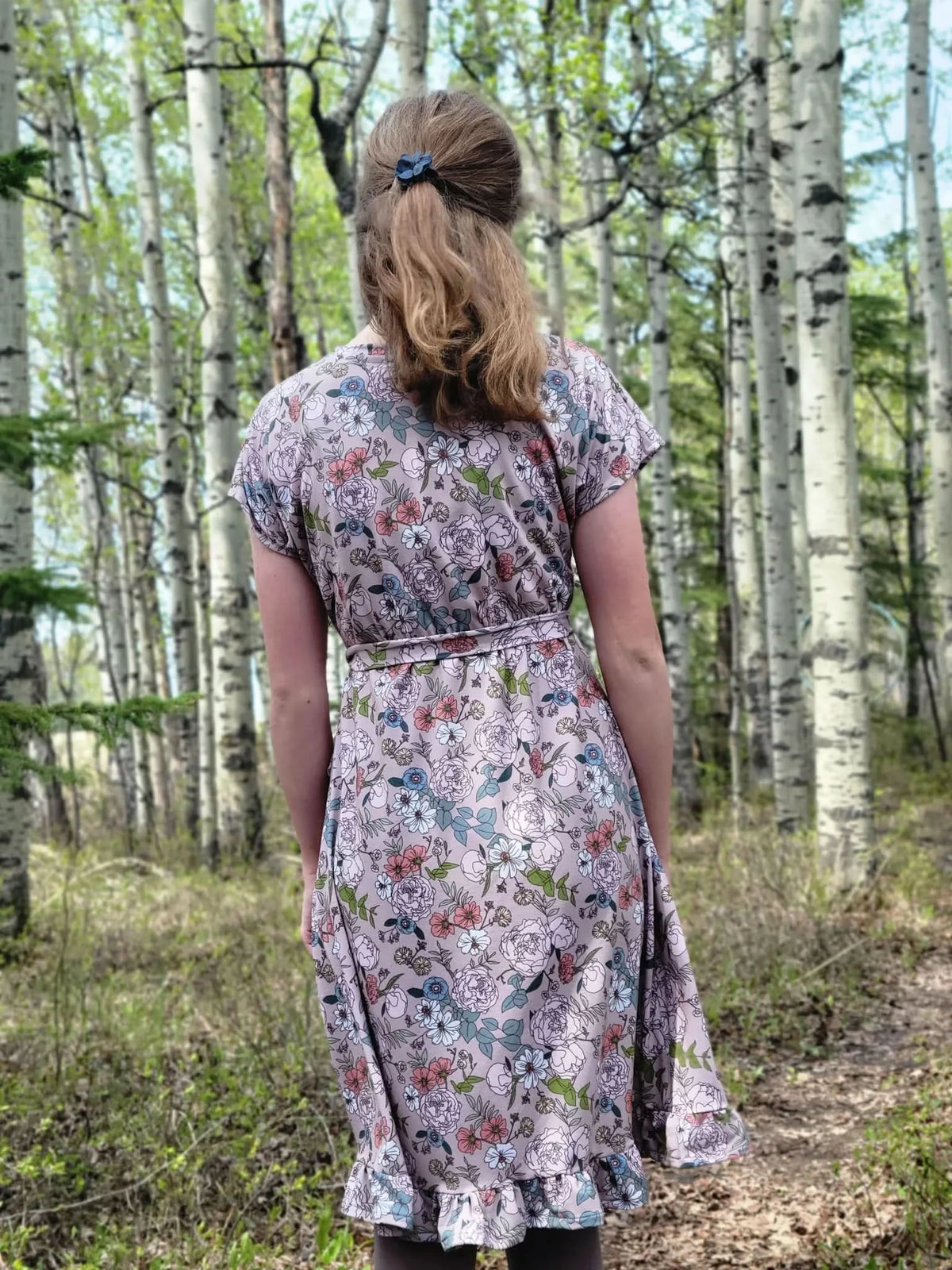A woman from behind wearing a tan floral dress in a forest.