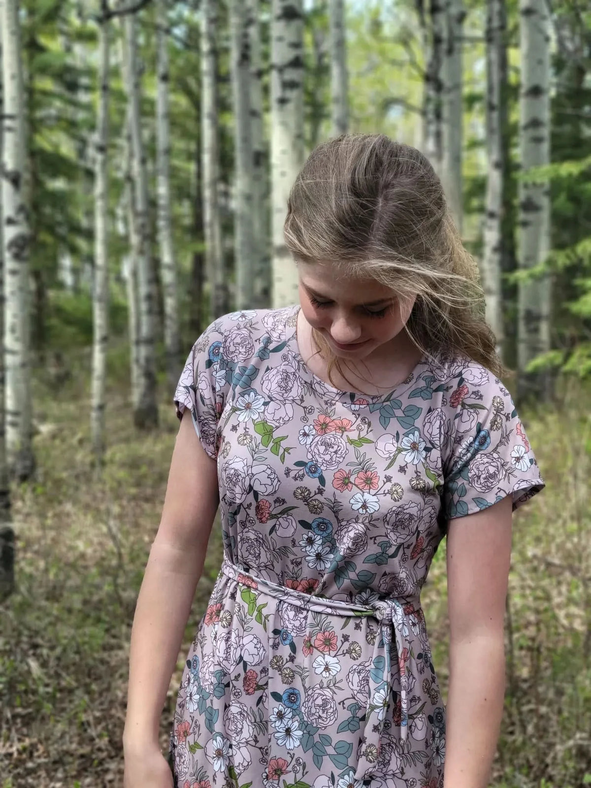 A young woman in a tan floral dress with a tie waist looks down in a forest.