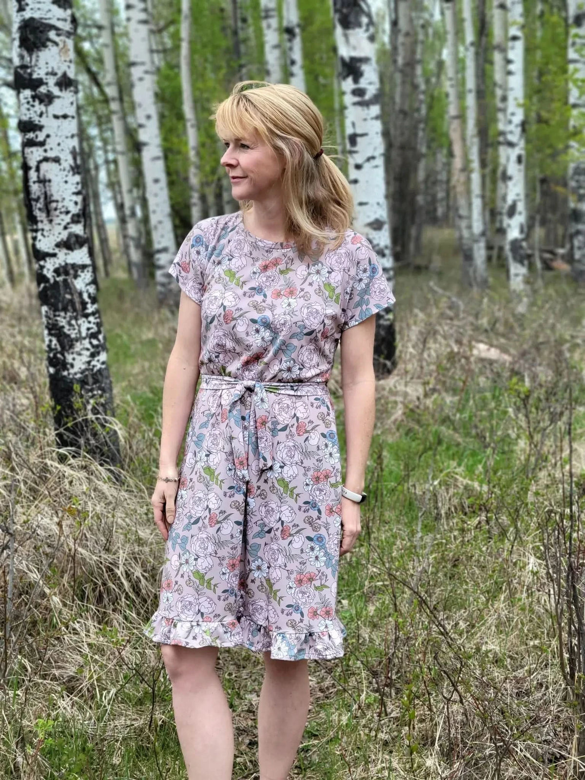 A woman in a floral dress stands in a forest with birch trees.