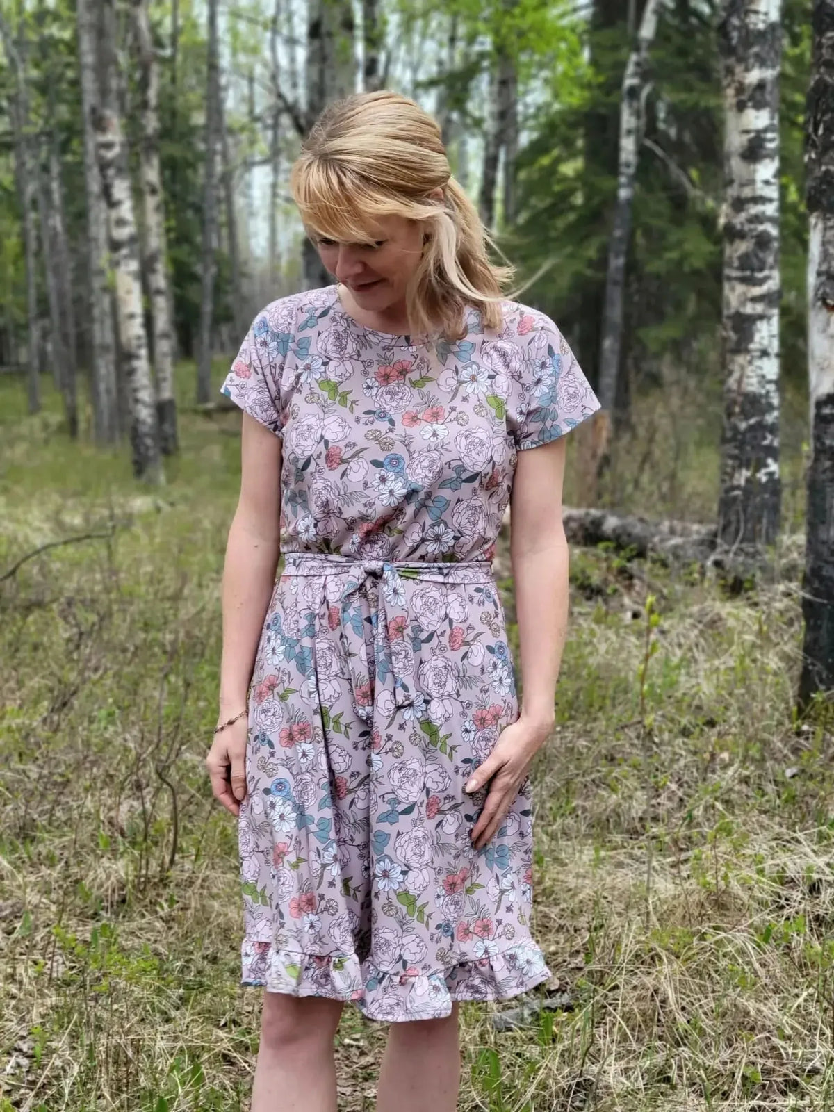 A woman in a tan Japanese floral dress with a ruffled hem stands in a forest.