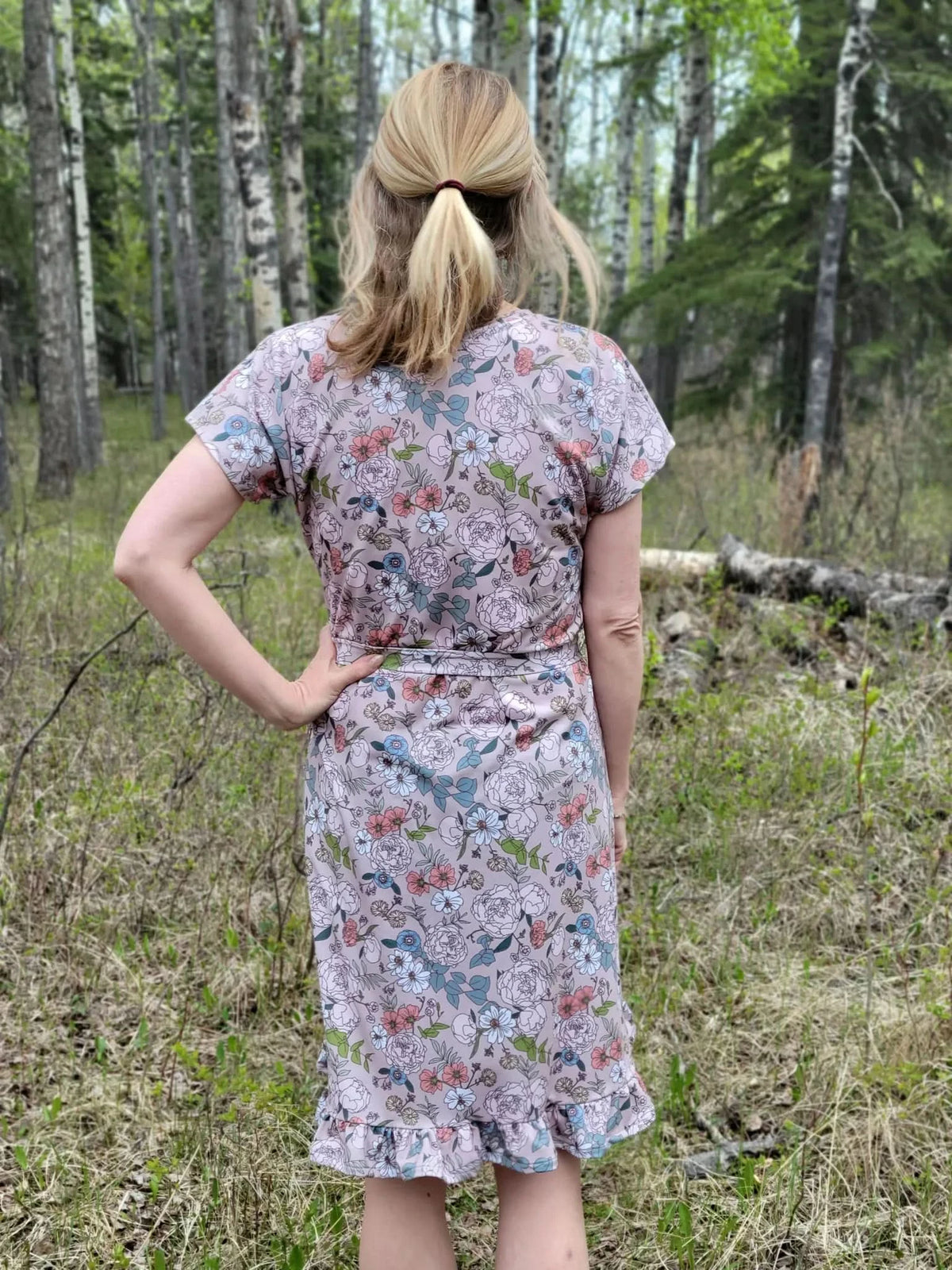 A woman from behind wears a tan floral dress with a ruffled hem in a forest.