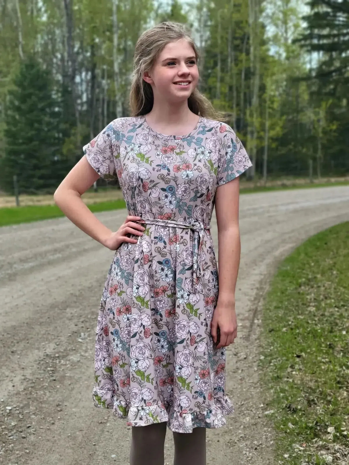 A young woman in a tan floral dress with a ruffled hem stands on a dirt road.