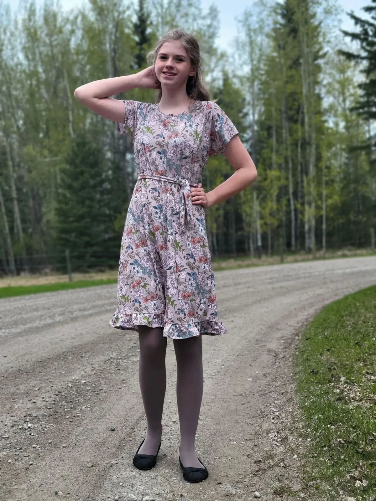 A young woman in a tan floral dress with a tie waist stands on a dirt road.
