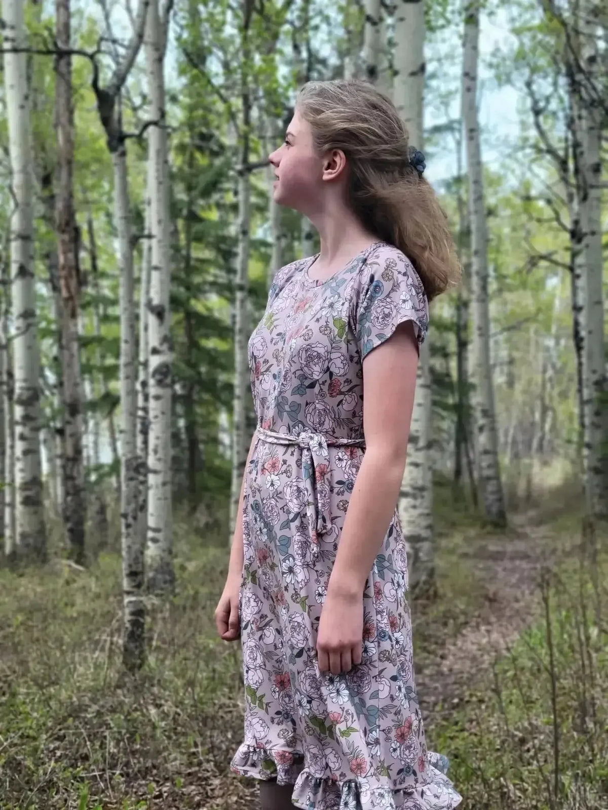 Side profile of a teen girl in a short-sleeved, belted tan floral dress amid birch trees.
