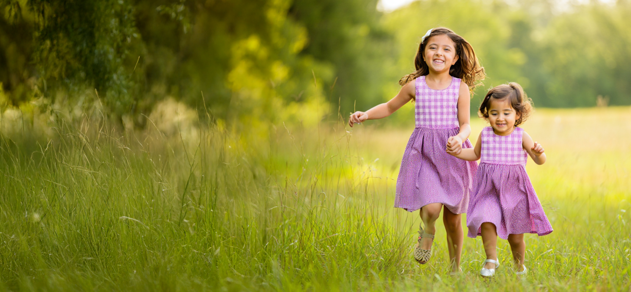 Two young girls in matching purple dresses walking through a grassy field.